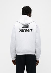 Άτομο με κοντά σγουρά μαλλιά φοράει ανοιχτό γκρι φούτερ με μαύρο λογότυπο «bareen» και μαύρο παντελόνι, κοιτάζοντας προς τα πίσω σε απλό λευκό φόντο.