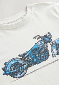 Balta kokvilnas t-krekls ar zilu motocikla grafiku, kurā ir izcila līniju zīmēšana, riteņi un stūres pret vienkāršu fonu.