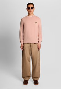 Άνδρας που φοράει σκούρα γυαλιά ηλίου, ροζ πουλόβερ με στρογγυλή λαιμόκοψη και μικρό κίτρινο λογότυπο, φαρδιά μπεζ παντελόνια και καφέ παπούτσια, στέκεται μπροστά σε γκρι φόντο.