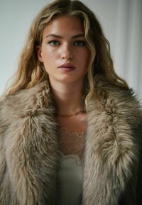 Faux fur frakke i lysebrune nuancer med en tyk, blød tekstur. Har en bred krave og lagdelt, flydende design over en blondetop.