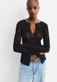 Schwarzes Spitzenoberteil mit langen Ärmeln und Rundhalsausschnitt, das vorne mit einem Schlüssellochdetail versehen ist. Wird über hellblauen Jeans und einem braunen Gürtel getragen.