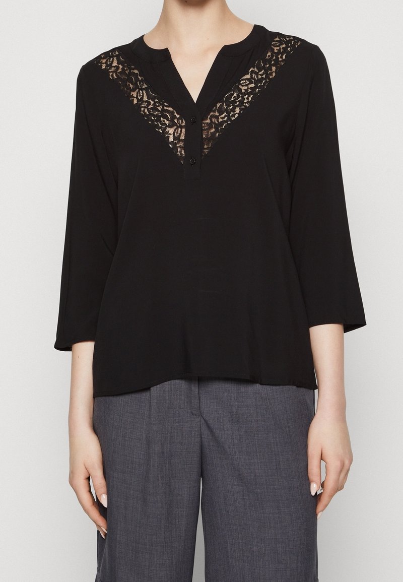 Frau in schwarzer Bluse mit langen Ärmeln und Spitzen-V-Ausschnitt sowie grauen weiten Hosen, stehend vor einem neutralen Hintergrund.