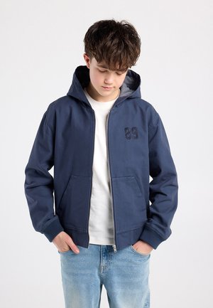 Veste zippée bleu marine avec capuche, doublure grise et numéro "89" brodé sur la poitrine. Deux poches avant et poignets côtelés.