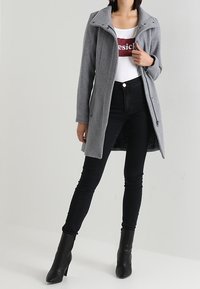 Cappotto grigio in misto lana con colletto alto, zip frontale e tasche laterali, indossato sopra una maglietta bianca e jeans neri aderenti con stivaletti.