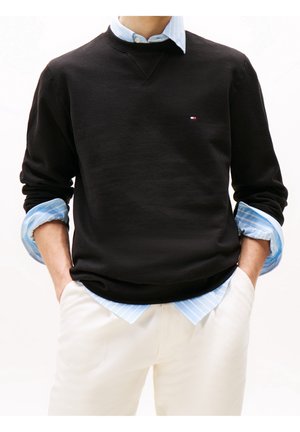 Person trägt einen schwarzen Pullover über einem blau gestreiften Hemd, weiße Hose, Hände in den Taschen, kleines Logo auf der Brust des Pullovers.