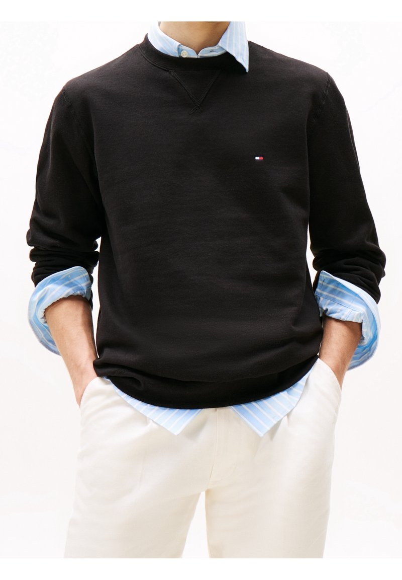 Personne portant un pull noir sur une chemise à rayures bleues avec un pantalon blanc, mains dans les poches, petit logo sur la poitrine du pull.