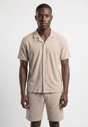Homme portant une chemise beige à manches courtes boutonnée et un short assorti, debout devant un fond blanc uni.