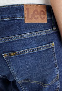 Buzunar din spate al blugilor albaștri din denim, cu cusături galbene și etichetă de piele maro a brandului Lee pe talie și o mică etichetă din material pe buzunar.