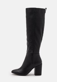 Botas negras de piel de cocodrilo hasta la rodilla con punta afilada, parte superior texturizada y tacón grueso. Incluyen una cremallera para ponerlas y quitarlas fácilmente.