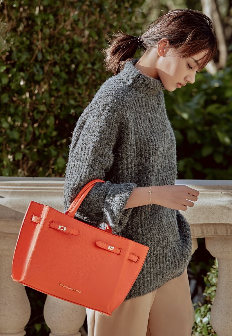 Orange läder tote-väska med kraftiga handtag, slät textur och silverdetaljer. Bärs med en grå stickad tröja och beige byxor.