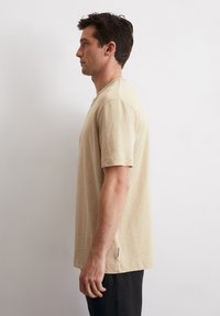 Beige T-Shirt mit kurzen Ärmeln aus strukturiertem Stoff, ausgestattet mit einem Rundhalsausschnitt und einem kleinen Seitenschild. Gerader Schnitt und entspannte Passform.