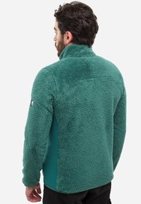 Mann, der sich mit dem Rücken zur Kamera dreht und eine türkisgrüne Fleecejacke mit hohem Kragen und strukturiertem Material sowie schwarze Hose trägt, vor einem schlichten Hintergrund.