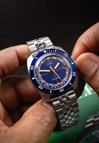 Une main ajuste une montre bracelet Timex en métal argenté avec un cadran bleu et des détails orange et blancs, sur un fond sombre.