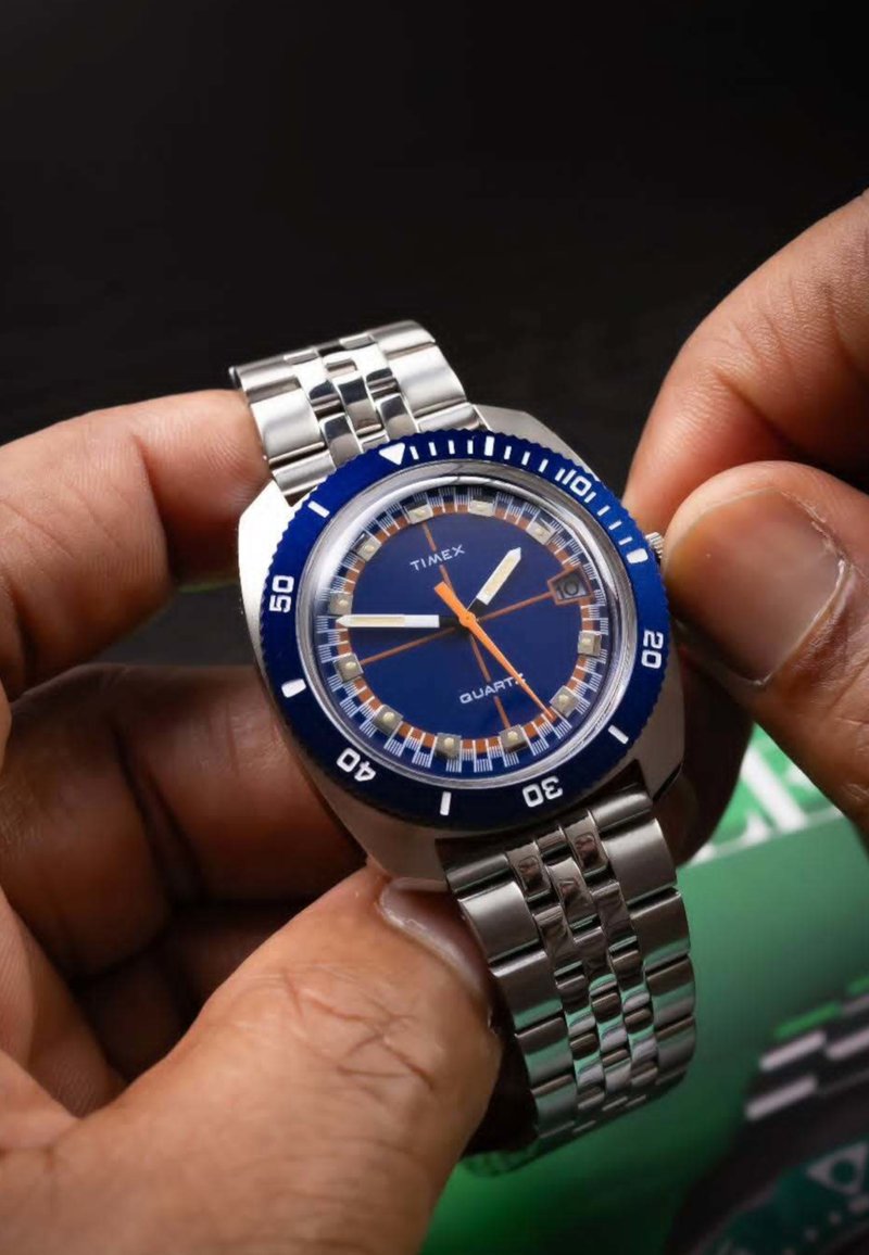 Une main ajuste une montre bracelet Timex en métal argenté avec un cadran bleu et des détails orange et blancs, sur un fond sombre.