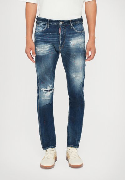 Dunkelblaue Denim-Jeans im Slim-Fit-Schnitt, mit Distressed-Details und subtilen roten Farbspritzern. Getragen mit schneeweißen Sneakern.