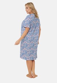 Kobieta stojąca boso, ubrana w niebieską sukienkę do kolan z krótkim rękawem w różowo-białe kwiatowe wzory, zwrócona bokiem z głową obróconą w tył.