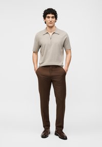 Bărbat stând cu mâinile în buzunare, purtând un polo bej cu mâneci scurte și fermoar, pantaloni maro și pantofi eleganți maro închis pe fundal alb.