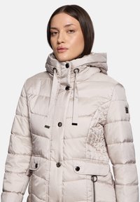 Veste matelassée beige clair avec capuche, dotée d'un design matelassé, fermetures à glissière et boutons, et deux poches avant avec des accents noirs.