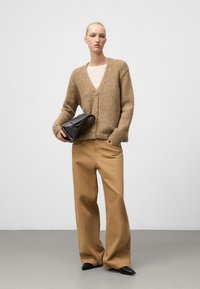 Brązowy sweter z dzianiny z głębokim dekoltem w serek, luźny krój i dużymi guzikami; w zestawie z beżowymi spodniami o szerokich nogawkach i małą czarną torebką.