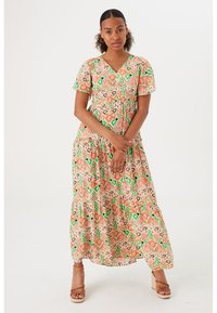 Mujer de pie con un maxi vestido de estampado colorido y mangas cortas, junto con sandalias de tacón marrón, contra un fondo blanco liso.