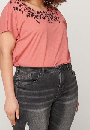 Femme portant un chemisier rose avec un imprimé de feuilles noires près de l'encolure et un jean noir délavé avec des légères usures sur la cuisse gauche.