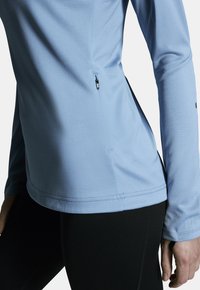 Hellblaues, langärmliges Sportoberteil mit seitlicher Reißverschlusstasche und strukturiertem Stoff. Es zeichnet sich durch ein tailliertes Design und glatte Nähte aus.