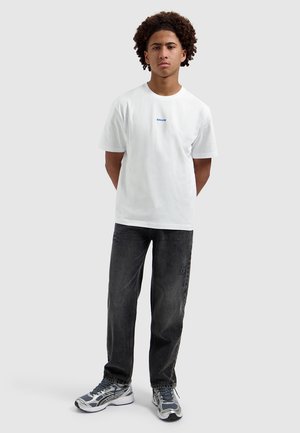 Young man with curly hair stands facing forward wearing a white T-shirt, black jeans, and gray sneakers against a plain background.