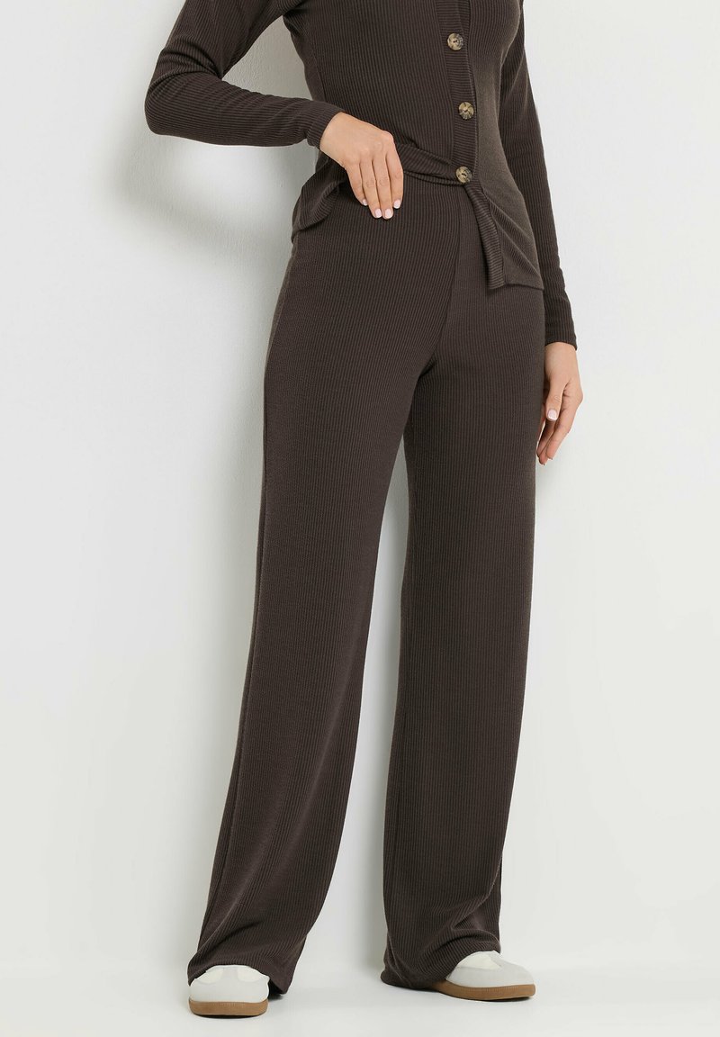 Person wearing ribbed dark brown wide-leg pants, matching button-up top, and white slip-on shoes standing against a white wall.