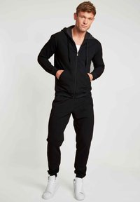 Zwarte zip-up hoodie met rode accenten en zakken, gecombineerd met bijpassende zwarte joggers. Witte sneakers maken de sportieve look af.