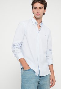 Ριγέ πουκάμισο με κουμπιά σε ανοιχτό μπλε και λευκό, χαρακτηριστικά με γιακά και λογότυπο στο στήθος, σε συνδυασμό με ανοιχτό τζιν παντελόνι. Λείο ύφασμα.