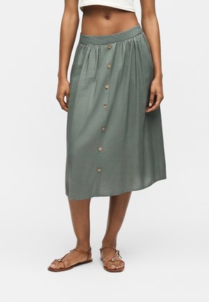 Femme portant une jupe vert olive longueur genou avec des boutons sur le devant et des sandales plates marron, debout devant un fond blanc.
