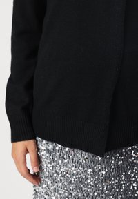 Person som bär en svart långärmad tröja och en silverfärgad paljettkjol, som visar en nära bild av överkroppen och vänsterhanden.