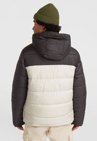 Zwarte en crèmekleurige pufferjack met een capuchon, met geborduurde secties. Het model draagt een groene gebreide beanie. De stof lijkt glanzend en gestructureerd.
