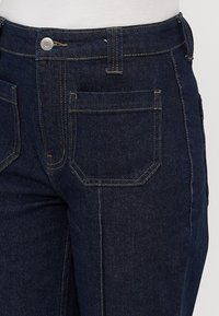Närbild av mörkblå denimjeans i midje- och övre lårområde med synlig framficka, knapp och sömnadsdetaljer.