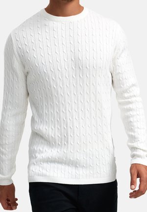 Homme portant un pull blanc à manches longues en maille torsadée avec poignets et ourlet côtelés, associé à un pantalon noir, sur un fond uni.