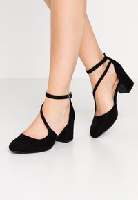 Zapatos negros de tacón con lados abiertos y correas en el tobillo, llevados en los pies de un modelo, de pie sobre una superficie brillante.