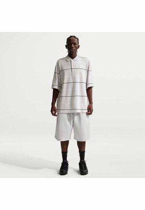 Persona in piedi che indossa una polo grigio chiaro oversize con sottili strisce nere, pantaloncini bianchi al ginocchio, calzini neri e sneakers nere.