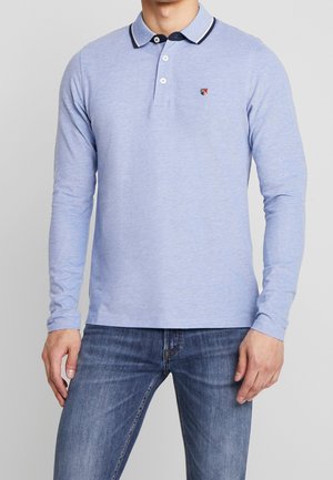Homme portant un polo bleu clair à manches longues avec col et trois boutons, associé à un jean bleu moyen sur fond uni.