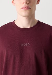 Burgunderfarbendes Crew-Neck-T-Shirt aus weicher Baumwolle mit einem erhabenen "BOSS"-Logo in passender Farbe auf der Brust. Glatte Textur.