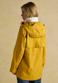 Geltona vandeniui atspari striukė su gobtuvu, balta apykakle, nugaros ventiliacijos detale ir ilgomis rankovėmis, nusistatanti neutralioje fone.