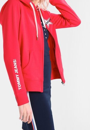 Osoba nosząca czerwoną bluzę z kapturem na zamek z napisem „TOMMY JEANS!” na rękawie oraz granatową sukienkę z bocznymi paskami w kolorze białym, czerwonym i niebieskim.