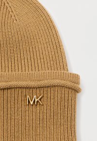 Bonnet en tricot marron avec une texture côtelée et une bordure repliée. Présente un accent de logo métallique doré "MK" sur le côté.