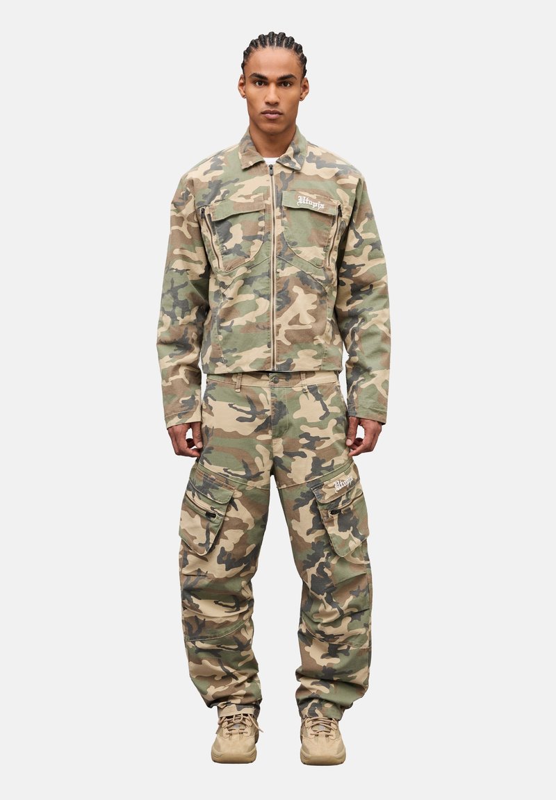 Uomo con capelli intrecciati che indossa una giacca mimetica con cerniera e pantaloni cargo con molteplici tasche, in piedi di fronte a uno sfondo bianco.