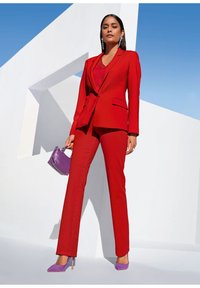 Roter maßgeschneiderter Anzug mit einer taillierten Blazerjacke und passender Hose. Lila Handtasche und Absätze. Klarer blauer Himmel im Hintergrund mit geometrischen weißen Formen.