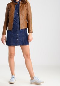 Femme portant une veste en cuir marron, une robe en denim bleu à boutons, et des baskets bleu clair, se tenant devant un fond clair.