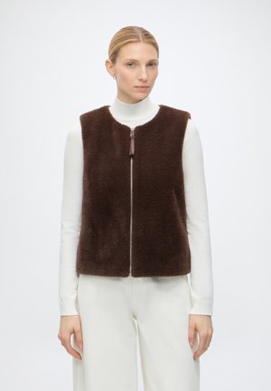 Frau trägt eine braune, flauschige Weste mit Reißverschluss über einem weißen Langarm-Rollkragenpullover und weißen Hosen, steht vor einem einfarbig hellen Hintergrund.