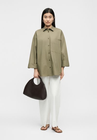 Γυναίκα που φοράει υπερμεγέθη πουκάμισο σε ελιά πράσινο, λευκό παντελόνι με φαρδιά μπατζάκια και τελείωμα με τρυπητό σχέδιο, καφέ σανδάλια, κρατώντας μεγάλη σκούρα καφέ τσάντα χειρός.