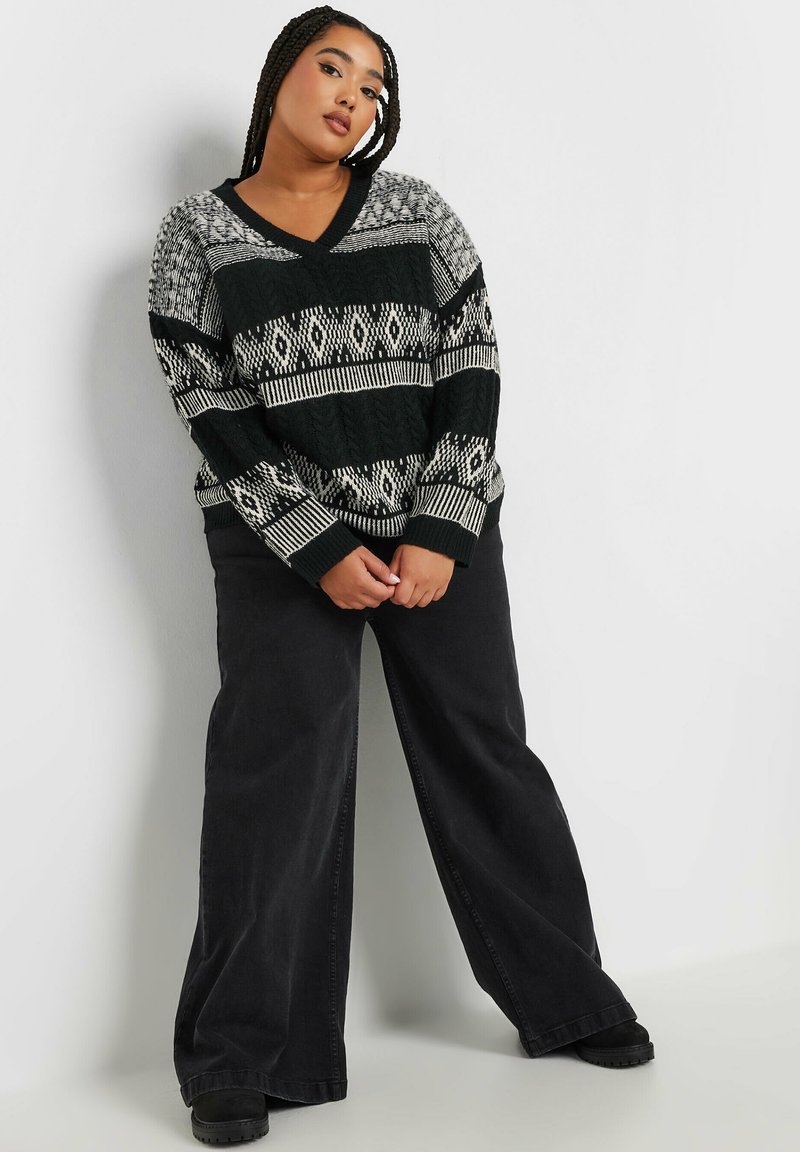 Strickpullover in Schwarz und Creme, mit V-Ausschnitt, geometrischen Mustern und gerippten Bündchen. Kombiniert mit locker sitzenden schwarzen Hosen.