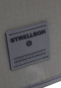 Tessuto grigio con un motivo a griglia presenta una toppa rettangolare nera con il logo STRELLSON e testo in rilievo, creando un contrasto di texture.