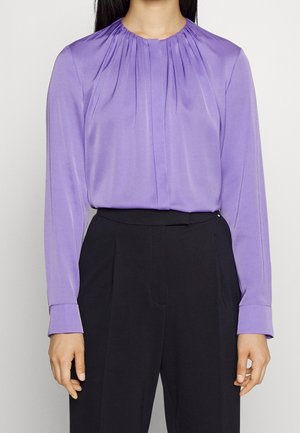 Vrouw met een lavendelkleurige, geplisseerde blouse met lange mouwen in een zwarte broek met hoge taille, getoond van schouders tot dijen.